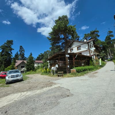 The image depicts a scenic view of a building complex and its surroundings in Atoluka, Bulgaria. A multi-story white building with brown trim and balconies is prominently featured, nestled amongst tall pine trees. Adjacent to this building is a wooden-framed structure with a glass-enclosed porch, likely a restaurant or cafe, adorned with a Bulgarian flag. In front of the porch, a white decorative horse statue stands on a wooden base, with a wooden bench nearby.

In the foreground, a silver SUV, identified by the license plate PA 7214BH, is parked on a gravel and dirt path. To the left of the SUV, a portion of a red vehicle is visible. The ground is a mix of dirt, gravel, and cracked asphalt, indicating a somewhat unpaved or weathered area. A paved road curves to the right, leading uphill and disappearing into the dense forest. The sky above is a clear, vibrant blue with scattered white cumulus clouds. The environment appears to be a mountain or forest resort area, with the buildings blending into the natural landscape. The scene is captured on a bright, sunny day.