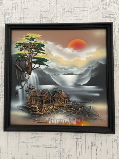 The image is a framed, three-dimensional painting featuring a landscape scene. In the foreground, two rustic, wooden houses are perched on a rocky outcrop dotted with gold glitter. To the left, a waterfall cascades down a cliff face, topped by a stylized tree with green and yellow foliage, also embellished with gold glitter. The middle ground depicts a body of water reflecting the sky, with dark, mountainous terrain in the background. The sky is rendered with a warm gradient from a reddish-orange sun to lighter tones, with stylized clouds. The painting is enclosed in a black frame. The artwork is hung on a textured, light-colored wall. There are no people or animals present in the scene. The location in Альхоўскі сельскі Савет, Belarus, is not visually represented in the painting itself; it is external context provided about where the image was taken.