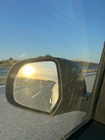 A rearview mirror of a car shows a bright sun on the horizon, reflecting on the glass which is speckled with dust. The mirror is mounted on the exterior of the car, with a road and a barren landscape visible in the reflection and outside the car. A small insect is perched on the casing of the rearview mirror. The sky is clear and blue. The location is Aleksandrovo, Bulgaria.