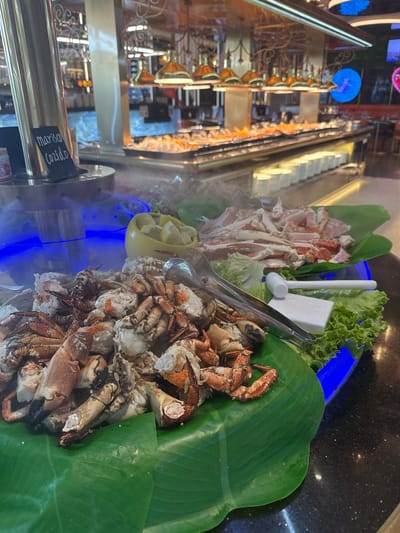 This media file shows a seafood buffet in what appears to be a restaurant, with a focus on crab. The foreground features a large pile of cooked crab legs and claws, artfully arranged on large green leaves, likely banana leaves, as a base. Next to the crab, there are pieces of white cheese and some lettuce. In the background, a buffet serving area is visible, with a stainless steel dispenser and some blue lighting. Further in the background, escalators and store displays suggest a shopping mall or a large establishment. The overall impression is a well-presented food display. The label on the dispenser, partially visible, reads "Marisco Cozido," which translates to "Cooked Seafood" in Portuguese. The lighting and steam rising from the dispenser suggest the food is freshly prepared and warm. No people are clearly visible in the frame. The setting appears to be indoors, with artificial lighting. Given the label and the general presentation, the location is likely a restaurant or a hotel buffet in Amora, Portugal.