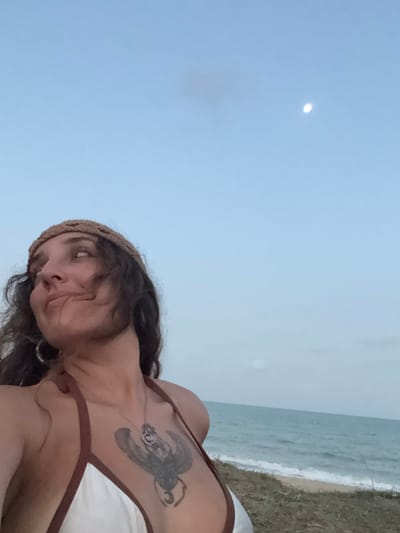 The image depicts an adult female on a beach in Ban Pang Pring, Thailand, during twilight or dawn. She is positioned in the left-central foreground, looking upwards and to the right with a slight smile. She has dark, wavy hair and wears a light-colored bikini top with brown trim, complemented by a light brown crocheted headband and a silver hoop earring on her left ear. A prominent dark-inked tattoo of a winged scarab or insect is visible on her sternum. The background features the ocean with small waves on a narrow shoreline, under a pale blue sky where a distinct crescent moon is visible in the upper right.