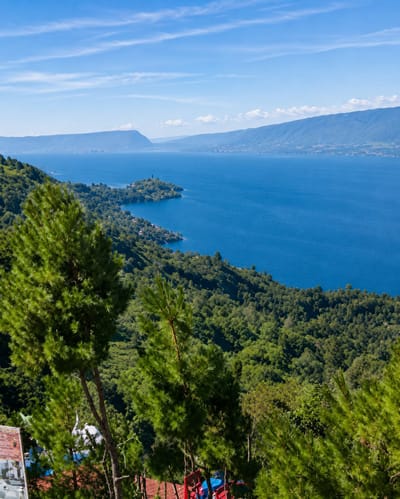 This is a wide-angle, eye-level shot of a serene landscape in Parik Sabungan, Indonesia, featuring a vast, blue lake with a backdrop of lush, green mountains under a clear, bright sky. In the foreground, several tall pine trees with vibrant green needles partially obscure the view of the lake and mountains. The trees are interspersed with glimpses of a red-tiled roof and some partially visible structures, possibly indicating human habitation or recreational facilities. The lake, a prominent feature, is a deep blue, reflecting the clear sky and stretching towards the horizon where it meets the distant, hazy mountains. The mountainous terrain surrounding the lake is covered in dense, green forestation, giving the scene a natural and tranquil feel. The sky is a brilliant blue, with a few wispy white clouds adding texture. The overall impression is one of natural beauty and peacefulness, with a hint of human presence in the foreground.