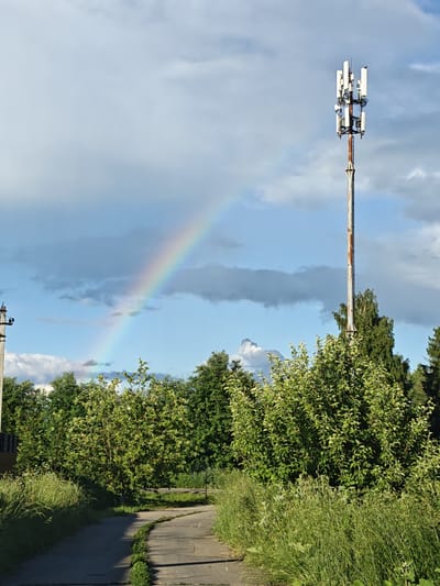 The image depicts an outdoor scene in Надежда Снт, Russia. In the foreground, a winding concrete path is bordered by tall, green grass. A dense cluster of leafy green trees and bushes dominates the middle ground. To the right, a tall, rust-colored cell tower stands against a cloudy sky, adorned with multiple antennas at its apex. A faint rainbow arcs across the sky behind the cell tower, originating from the left side of the frame. On the far left, a utility pole with electrical wires is visible. The environment appears to be a semi-rural or suburban area, likely during daylight hours with a mix of sun and clouds.