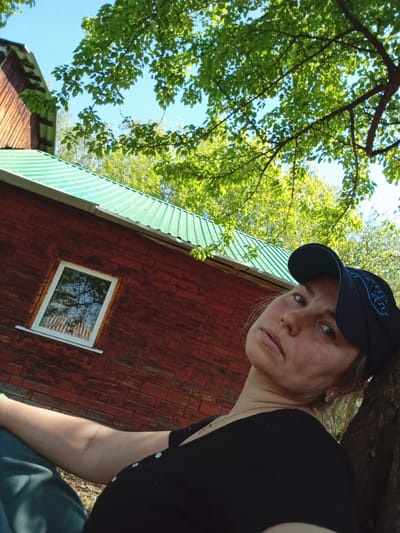 A woman in a black t-shirt and a dark blue baseball cap leans against a tree trunk. Behind her is a red wooden building with a green corrugated metal roof and a white-framed window reflecting trees. The sky is visible through the green foliage overhead. The scene appears to be outdoors during daylight. There is no verifiable evidence to confirm the location is Монастырка, Russia.