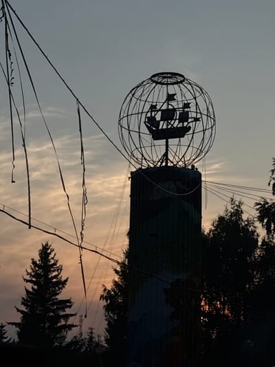 The image captures a striking silhouette of a cylindrical tower topped with a large, spherical metal cage containing the silhouette of a sailing ship. This structure, likely a monument or art installation, is located outdoors in Vekoshinka, Russia. The setting appears to be twilight, with a soft, orange and gray sky filled with wispy clouds. Silhouetted evergreen trees are visible in the foreground and to the right, framing the tower. Several thin wires or streamers hang from unseen points, creating a dynamic, almost chaotic, visual element against the sky. The tower itself is dark and appears to have some painted detail on its surface, which is difficult to discern in the low light. The overall impression is one of atmospheric beauty and possibly a touch of mystery, given the unusual nature of the monument. There are no people visible in the image.