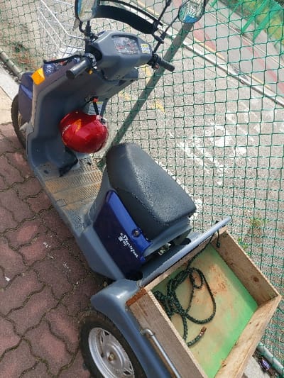 A gray and blue "Shevron" electric scooter (쉐브론 전기자전거 107) with a black seat is parked on a red-brown interlocking paver pathway in 강화군, South Korea. A bright red helmet hangs from the left handlebar, positioned near a front-mounted silver wire basket. The scooter features chrome handlebars, rearview mirrors, and a visible dashboard. Attached to the rear of the scooter is a rectangular wooden cargo cart with weathered light green base material, containing a coiled dark green and black mottled rope. A green chain-link fence is visible behind the scooter, separating the paver pathway from a lighter-colored paved surface with yellow markings in the background.