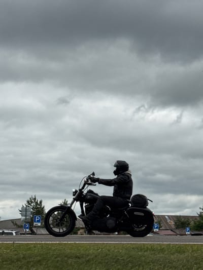 In Privolzhsky, Russia, a person wearing a helmet and dark clothing rides a black motorcycle on a paved road under a cloudy sky. The motorcycle has saddlebags and is in motion. In the background, there are buildings, trees, and parking signs. The scene appears to be on the outskirts of a town or in a rural area.
