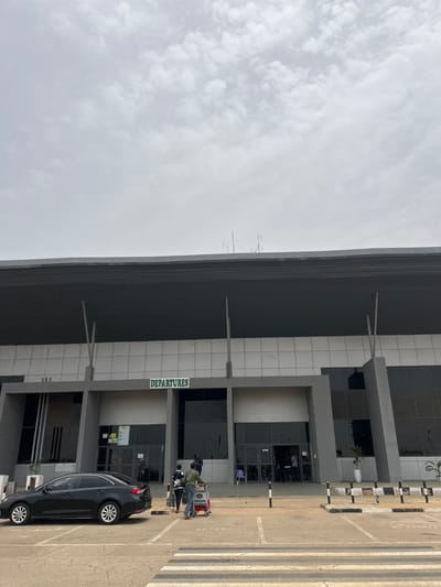 The scene depicts the exterior of an airport departures terminal in Kwi, Nigeria. The building is modern, with a gray and white facade, featuring a prominent "DEPARTURES" sign. Several people are present, interacting with luggage carts near the entrance. A black car is parked nearby. The sky is overcast. The area in front of the terminal features parking and pedestrian walkways.