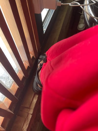 The image depicts a close-up, first-person perspective view of a person's right leg, primarily encased in bright red fabric, likely trousers. The foot is shod in a dark, multi-strapped open-toed sandal. To the left, a series of vertical wooden slats cast shadows across a light-colored, textured floor or ground. Beyond the slats, an exterior or window is suggested by a brighter area. In the background, to the right of the red leg, a white power adapter is plugged into an unseen outlet, with a white cable extending downwards. A white athletic shoe is partially visible on a dark floor in the mid-ground, and a portion of a dark, reflective object, possibly a helmet, is present above it. The scene is illuminated by strong natural light, creating defined shadows. The location is Gatongora ward, Kenya.