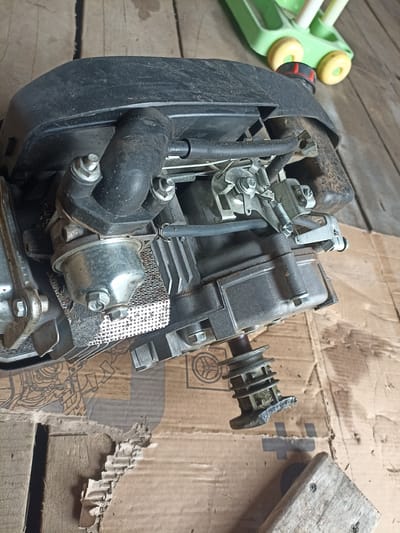 The media file displays a close-up, overhead view of a disassembled engine, likely from a lawnmower or similar equipment, resting on a wooden floor and cardboard. The engine is the primary object, with its black plastic cover, carburetor, and various metal components clearly visible. A portion of a green and yellow toy, possibly a ride-on toy with wheels, is visible in the upper right background, suggesting a domestic or storage setting. The overall scene is dimly lit, with no indication of specific weather conditions. The engine appears to be in a state of repair or maintenance, with its parts laid out. The background is rustic, with weathered wooden planks forming the floor. Some faint, abstract line art is visible on the cardboard beneath the engine. No people are present in the image. The focus is solely on the mechanical components of the engine.