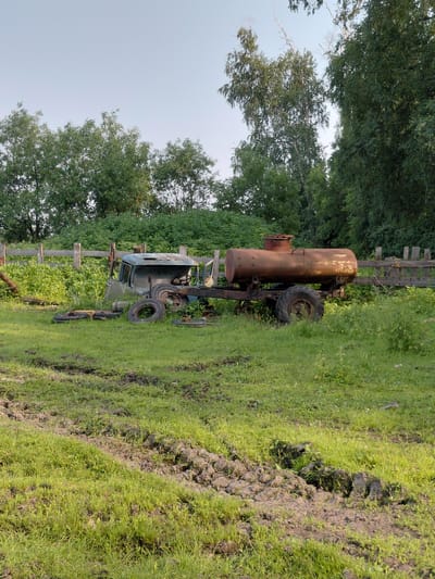 In a grassy, uneven field in Novoselovo, Russia, an abandoned, rusted water tank trailer is hitched to a derelict, light blue truck cab. The truck cab is missing its body and appears to be partially sunk into the muddy ground. Several loose tires are scattered around the truck. A wooden fence runs along the back of the property, with lush green trees and foliage behind it. The sky is clear and pale blue. The scene suggests a rural or semi-abandoned setting, possibly a farm or storage area, where old equipment has been left to decay.