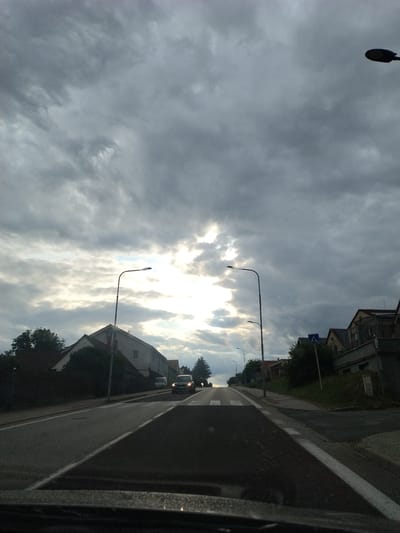 The image captures a road scene in Dražice, Czechia, under a cloudy sky with patches of sunlight breaking through. A paved road with visible white lane markings stretches into the distance, leading uphill. A pedestrian crossing is marked on the road. On the left side of the road, there are houses and trees. On the right side, there are more houses and a pedestrian crossing sign. A black car is visible on the road, heading towards the viewer. Streetlights are present on both sides of the road. The overall atmosphere is overcast but with some bright areas due to the sun.