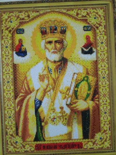The image is a diamond painting depicting Saint Nicholas. The artwork is framed in a golden border with intricate patterns. The central figure of Saint Nicholas is rendered in a mosaic of small, colored diamonds, creating a textured and shimmering effect. He wears a golden crown and ornate religious vestments, and holds a staff in one hand and what appears to be a book or scroll in the other. Flanking him are two smaller figures, one on each side, also depicted in diamond mosaic. The background is a radiant halo of golden diamonds, with architectural elements and decorative borders suggesting an icon. Below the central figure, a red band displays Cyrillic text. The diamond painting is likely a decorative item for a home or religious space. The location in the city of Hiliuți, Moldova, is not visually represented in the image itself; it is external information provided with the image.