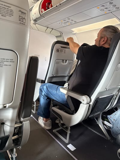 The image depicts the interior of a commercial aircraft cabin. The scene is reportedly situated within the context of AMCO, Area Metropolitana Centro Occidente, Colombia.
An adult male, with graying hair and a beard, occupies an aisle seat in the foreground-middle. He wears a black short-sleeved t-shirt, blue jeans, and light-colored athletic shoes with three dark stripes. His right arm is raised, with his hand resting on the top edge of the seatback directly in front of him. He is seated facing forward, leaning slightly. A gray seatbelt is visible across his lap.
The aircraft interior features white and light gray paneling, with dark gray/black seats. The seatbacks are equipped with tray tables and lower seat-back pockets. A "JetSMART" logo and instructional text in English ("under your seat") and Spanish ("salvavidas debajo") are visible on the seatback directly in front of the camera, to the left of the main subject. A QR code and additional text are visible on the seatback in front of the main subject.
Overhead, white storage compartments are present, one of which is partially open, revealing a red object inside. An overhead panel with circular light and vent controls is also visible. The floor is covered with a dark gray textured material, and a small, rectangular white object lies on the floor near the main subject's feet. Partial views of other passengers or their belongings are visible in the seats adjacent to and in front of the main subject.