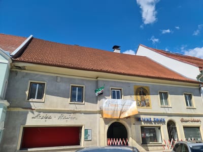 The image shows a building facade under a blue sky with scattered clouds in Sankt Marein bei Neumarkt, Austria. The building has a reddish-brown tiled roof and a light-colored stucco exterior. There are several windows on the upper floor, some with white frames and others with darker frames. A Syrian flag is hanging from one of the upper windows.

Below the windows, there is a large banner with text and a logo, reading "ET KÖNIG" and "www.et-koenig.at". The banner is orange and white. Further down on the facade, there are two shopfronts. The left shopfront has a large red covering or awning. The right shopfront has signage that appears to read "VZ VINZ. ZWICK" and "Eisen Waren". In front of the shops, there are barriers made of red and white striped plastic.

A fresco or mural depicting a man in historical attire holding a hammer and anvil is painted on the upper part of the facade to the right of the Syrian flag. There are also some inscriptions on the stucco above the shopfronts. The overall impression is of a commercial building in a town.
