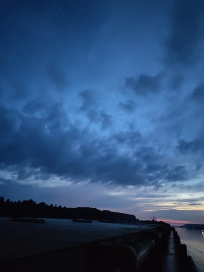A twilight scene unfolds over a dark body of water, likely a river or lake, in Badozhskoi Pogost, Russia. The sky is a deep, moody blue, filled with dramatic, dark clouds that obscure most of the fading daylight. A thin trail, possibly from an aircraft, is faintly visible against the cloud cover.
In the foreground, a dark, solid structure, possibly a pier or embankment, stretches along the lower right and bottom of the frame, its surface indistinct in the low light. To the left of this structure, a flat expanse of lighter ground meets the dark water. Beyond this, a silhouetted line of trees marks the distant shore.
Towards the right side of the image, the water stretches towards the horizon. A faint orange and pink glow emanates from the western sky, suggesting the sun has recently set. A few distant lights, possibly from a settlement or vessels, dot the far shoreline. There are no visible people or specific activities depicted in the scene. The overall impression is one of quiet, natural beauty at the edge of darkness.