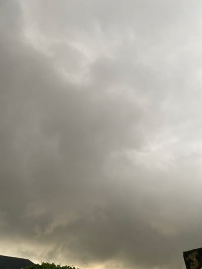 The image depicts a cloudy sky, with heavy grey clouds dominating the scene. In the foreground, at the bottom right, a portion of a concrete structure is visible, appearing weathered and possibly part of a building's exterior. To the left, the dark roof of a building is partially visible, and below it, green foliage suggests the presence of trees or plants. The lighting indicates an overcast day, likely before or during rainfall. No people are present in the image, and no specific actions or interactions are captured. The scene appears to be an outdoor environment in Jere South, Nigeria, characterized by atmospheric conditions indicative of impending or ongoing inclement weather.