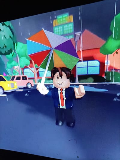 A Roblox avatar of a young person with dark hair, wearing a blue suit and red tie, stands in the rain holding a colorful umbrella. The avatar is in the foreground, facing the viewer with a surprised expression. Behind the avatar, a yellow car and a pink car are parked on a wet street, with puddles reflecting the surroundings. In the background, a red building with blue-paned windows and trees are visible. The scene appears to be set in a stylized, cartoonish city environment. There is no discernible connection to Alatyr, Russia.