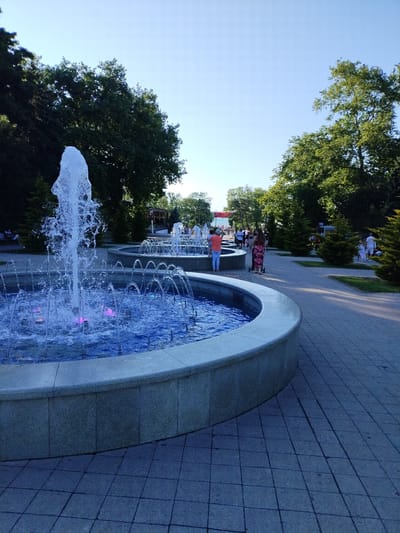 The image depicts a sunny day in a park with multiple fountains. The foreground features a large fountain with illuminated blue water and colorful lights. In the background, several other fountains are visible, along with lush green trees and a clear blue sky. People are seen walking along the paved pathway beside the fountains, some with strollers. The location is identified as Vinogradny, Russia.