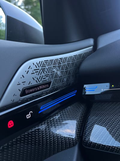 The image shows a close-up of a car's interior, focusing on the door panel and a section of the dashboard. A speaker grille with a triangular geometric pattern is visible, bearing the "Bowers & Wilkins" logo. Below this, there is a black panel with illuminated blue lines and icons, including a red locked padlock and a white unlocked padlock. To the right, another black panel features a digital display with a slider and a small green indicator light. The car's interior appears to be trimmed with carbon fiber material in the lower part of the frame. The background shows a blurred view of outdoor greenery, suggesting the car is parked outdoors. There are no people present in the image. The image does not contain any information that would suggest it is located in Robești, Romania.