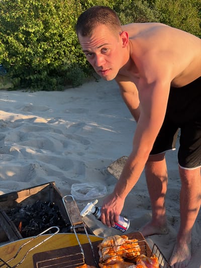 A shirtless man is bent over a grill on a sandy beach. He is holding a can of what appears to be lighter fluid and is spraying it towards the grill, which contains burning coals and raw chicken. The man has short brown hair and is wearing black shorts with white trim. The background consists of dense green foliage and sand. The location is the city of Povorovo, Russia.