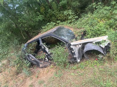 The image depicts the wreckage of a black car overgrown with vegetation. The car is heavily damaged, with its doors and hood open, and the front fender is detached. Numerous plants and vines have grown over and through the vehicle, obscuring much of its structure and suggesting it has been abandoned for a significant period. The setting appears to be a natural, overgrown area, possibly a roadside or a field, with trees and dense foliage in the background. The ground is covered in dry grass and green plants. The lighting suggests it is daytime, with natural light filtering through the trees. There is no visible text in the image. The scene evokes a sense of decay and nature reclaiming man-made objects. The location context provided is Lukovo, Bulgaria.