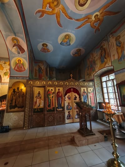 The image depicts the interior of an Orthodox church, specifically identified as being in the city of Kady, Russia. The scene is richly decorated with religious iconography, with numerous frescoes adorning the walls and ceiling. The ceiling features a blue expanse with painted depictions of angels with golden wings, and circular portraits of saints. The walls are covered in similar religious art, showing various figures in traditional Orthodox attire.

In the foreground, a prominent iconostasis, a wall of icons, separates the nave from the sanctuary. This structure is elaborately carved and painted, with multiple icons of saints and biblical scenes. To the right of the iconostasis, a wooden lectern stands, likely used for readings during services. In front of the lectern, a brass candelabra with lit candles illuminates the area.

The floor is tiled in a pattern of light and dark squares. To the left of the iconostasis, a large icon featuring a group of three figures in golden robes is visible. Further to the left, a wall with smaller framed pictures and a glimpse of what appears to be a candle holder suggests another area of the church. A window with intricate latticework is visible on the right side of the image, allowing natural light to enter the space. The overall impression is one of solemnity and religious devotion, characteristic of Orthodox Christian places of worship.