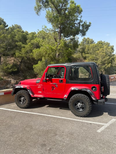 A bright red, two-door Jeep Wrangler Sport is parked on asphalt in an outdoor lot. The vehicle has a black hardtop and black fender flares that appear to have a distressed, faded camouflage pattern. A spare tire is mounted on the rear door. The Jeep has decals on its side, including a graphic of a Native American man with feathers and a smaller "Jeep" logo. The vehicle is angled towards the right of the frame. Behind the Jeep, a chain-link fence runs along a hillside covered in dense green pine trees. The sky is clear and blue. The lot is marked with white parking lines. A short, segmented white and red barrier is visible in the foreground on the left. The scene appears to be in a rural or semi-rural area. It is daytime, with bright sunlight casting shadows. The image was taken in the city of Gènova, Spain.