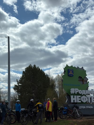 The image depicts a group of cyclists standing next to a roadside sign with the inscription "#РоднойНЕФТЕ... Душа Урала" and "#террабашкирия" in Vorob'evo, Russia. The sign is shaped like the outline of a green region with a coat of arms on it. There are about seven cyclists visible, each with a bicycle, and they appear to be resting or taking a break. The sky is cloudy, with patches of blue visible. The background includes trees and a grassy area. A utility pole is visible in the lower part of the image.