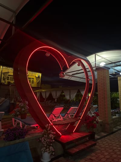 The image depicts an outdoor area at night, illuminated by neon lights and ambient building lights. The central focus is a large, red heart-shaped structure outlined with red neon, featuring two small disco balls hanging inside. In front of the heart, there is a wooden platform with steps. The area appears to be a recreational space, possibly a resort or a public park, with multiple lounge chairs visible. In the background, there is a building with lit windows and decorative string lights. To the right, a brick pillar supports a white canopy. Potted flowers and lush greenery are scattered throughout the scene, adding color and life to the nighttime ambiance. The paving stones on the ground suggest an urban or developed outdoor setting, possibly within the city of Yaroslavskaya, Russia. There are no people visible in the image.