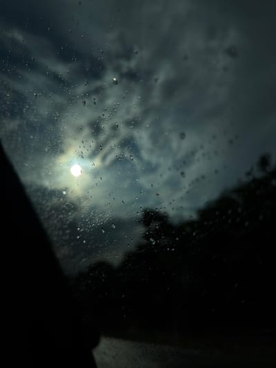The image depicts a view through a rain-streaked window, likely from inside a vehicle. The primary subject is the sky, characterized by dramatic, dark, and cloudy formations. A bright, luminous sun is visible through the clouds, creating a somewhat diffused and hazy glow. Numerous water droplets of varying sizes are scattered across the windowpane, distorting the view and adding a textured, abstract quality to the scene. The droplets catch the light, creating small, circular highlights.

In the upper right portion of the image, a dark, silhouetted mass suggests the presence of trees or foliage outside, further obscuring the sky. A dark, angled shape in the lower right corner could be the interior of the vehicle, such as the dashboard or a pillar.

The overall mood is somber and perhaps a little melancholic, evoked by the dark clouds and the rainy conditions. The bright sun peeking through suggests a break in the weather or a moment of contrast. The time of day is not explicitly indicated, but the quality of light could suggest late afternoon or a period of overcast skies with intermittent sunshine.

There is no visible text in the image. The setting is likely rural or suburban, given the hint of trees and the open sky. The presence of rain indicates a wet weather event.