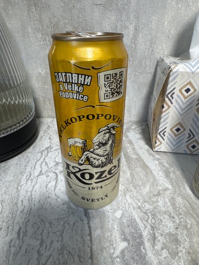 This is a medium close-up, rotating shot of a golden beer can. The can is oriented horizontally and rests on a textured, gray surface. Above the can, partially visible, is a box with a patterned design of beige, blue, and gray diamonds. Below the can, a ribbed, translucent object with black accents is visible.

The can features prominent branding. On the left side, in Cyrillic script, are the words "ЗАГЛЯНИ" and "В Velké Popovice". This text appears to be a call to action, perhaps "Look inside" or "Discover". To the right of this, there's a QR code. Further along the can, a black and white illustration depicts a goat-like creature holding a frothy beer mug, with the word "KOZE" emblazoned in large, stylized letters. The year "1874" is visible beneath "KOZE", and to the right, the word "SVĚTLÝ" is written vertically. The lighting suggests an indoor setting with ambient light, and no specific time of day or weather conditions are discernible. The overall impression is of a product display.