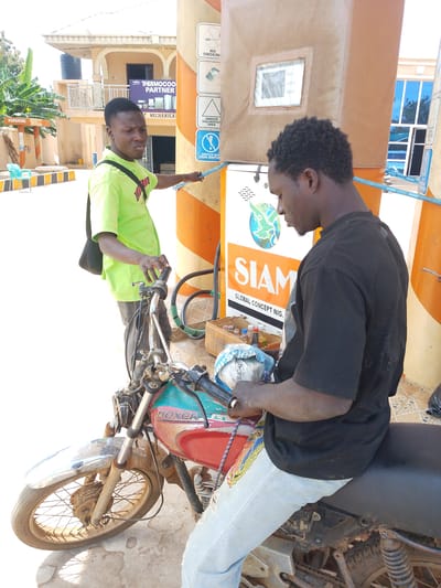 Two young men are at a gas station in Oke Iho, Nigeria. The man on the left, wearing a lime green t-shirt and a black shoulder bag, is standing and pointing towards a gas pump. He is holding a blue rope in his right hand. The man on the right, wearing a black t-shirt and light blue jeans, is sitting on a red and green motorcycle. The motorcycle's front wheel is visible, and it appears to be a Boxer model. He is holding a white cloth with some items wrapped inside. In the background, a building with a blue-tinted window is visible, along with a gas pump with various warning signs. The ground is paved with tiles.