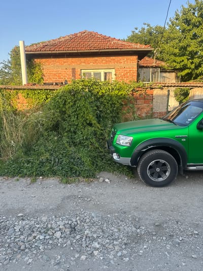 A green Ford Ranger pickup truck is parked on a gravel road next to a weathered brick house. The house has a red tile roof and is partially covered in lush green ivy. There is a concrete wall in front of the house, also overgrown with vegetation. The sky is clear and blue, indicating a sunny day. The scene appears to be in a rural or semi-rural setting, likely in Mednikarovo, Bulgaria, given the architecture and vegetation. There are no people visible in the image.