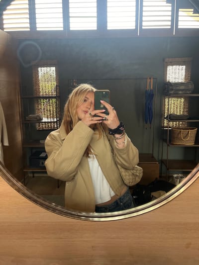 A person is taking a selfie in a round mirror in a room in Livingstone, Zambia. The person, with blonde hair, is wearing a beige jacket, a white shirt, and blue jeans. The room features a window with wooden slats, a shelf with baskets and umbrellas, and a small safe. The mirror is framed with wood.