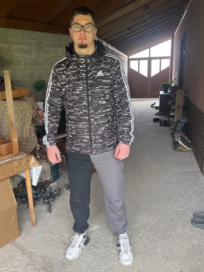 A male individual is standing in what appears to be a cluttered garage or storage area. He is wearing glasses, a black and white patterned Adidas jacket, one leg of gray pants and the other of black, and white sneakers. He is facing the camera. The background includes various items such as tools, boxes, plants, and miscellaneous objects, indicating a cluttered storage space. There are doors or a large opening in the background allowing light into the space. The location is unknown.