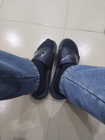 The image shows a first-person perspective of a person's legs and feet. The person is wearing blue denim jeans and black socks with black Nike slide sandals. The sandals have a white Nike swoosh logo on the strap of each sandal. The person's feet are resting on a beige tiled floor. The lighting suggests an indoor environment. The specific location in Abeere, Nigeria, is not discernible from the image itself.