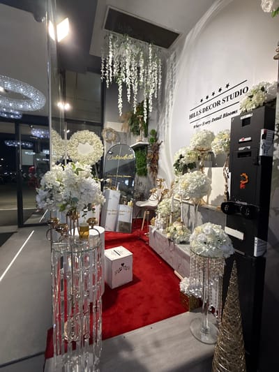 The image depicts the interior of a store, possibly a wedding or event decor studio, with a prominent sign on the wall reading "HILLS DECOR STUDIO Where Every Detail Blooms." The store is filled with white floral arrangements, crystal chandeliers, and decorative items, set against a white wall and a red carpet. In the foreground, a clear glass stand with hanging crystals holds a bouquet of white flowers. To the right, a tall black cabinet with several compartments is visible, and next to it is a golden, cone-shaped decorative object. The store appears to be located in Ilidža, Bosnia and Herzegovina, as suggested by a reflective surface displaying the word "Dobrodošli" (Welcome in Bosnian) and text referencing "Goran's 50th Birthday Celebration." No people are present in the image.