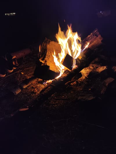 A campfire burns brightly at night in what appears to be a rural or undeveloped area, possibly near Kul'metovo, Russia, given the context. The fire is the central focus, with flames leaping upwards from a pile of logs and embers. The surrounding ground is covered in dark soil, dry grass, and twigs, illuminated by the firelight. In the foreground, to the left, a pair of dark sneakers are visible, suggesting the presence of people. The background is dark and indistinct, with a faint glow in the distance that could be distant lights. The overall scene evokes a sense of warmth and outdoor activity in a natural setting.