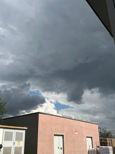The image captures an overcast sky with dramatic, dark storm clouds dominating the frame. A patch of bright blue sky is visible through a gap in the clouds, suggesting that the storm is either approaching or receding. To the right of the frame, a modern building with a pale pink facade is visible, featuring two white doors or windows. A small, red and white sign is affixed to the pink wall. Below the pink section of the building, a dark gray lower section is visible, with a white utility box featuring horizontal slats and a yellow warning label. In the background, stacks of what appear to be wooden pallets and white wrapped objects are piled high, along with a hint of green foliage. The overall scene suggests a building site or industrial area. The time of day is difficult to ascertain precisely due to the heavy cloud cover, but the brightness of the blue sky patch indicates it is daytime. The weather is clearly stormy or about to be. The location context is Tenevo, Bulgaria.