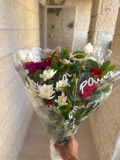 A male's right hand, adorned with a gold-colored chain bracelet, holds a mixed floral bouquet wrapped in clear plastic. The plastic bears white script lettering, notably featuring the words "power", "joyful", and "ordinary". The bouquet includes white chrysanthemums, dark pink dahlia-type flowers, red cockscomb, white eustoma, small red berries, a white rose, green eucalyptus foliage, and assorted filler flowers in white and brown tones. The background consists of a textured, light-colored stone wall. A blurry reflective surface, consistent with a glass door or window framed in a light color, is visible in the mid-ground, reflecting the stone wall. The scene is illuminated by natural light in Maale Adumim, Palestinian Territory.