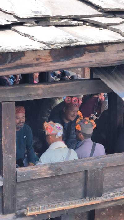 A medium-shot, eye-level, vertical image shows a group of men in traditional Himachali hats standing on a wooden balcony in Malana, India, likely during a festival. The men are gathered close together and appear to be engaged in conversation. The man on the far left is wearing a red and black patterned cap, a dark green long-sleeved shirt, and has a beard. To his right, a man with a red hat adorned with circular, dark red embellishments and bright pink accents is smiling and looking to his left. In the foreground, a man in a light-colored garment and a white hat decorated with colorful pompoms and sequins is facing away from the camera, looking towards the man in the red hat. Behind him, another man wears a grey cap with orange floral decorations and a light purple shirt. The background is dark, suggesting an interior space or a shaded area. The balcony is constructed of rough-hewn wooden planks, and above them, the distinctive slate roofs of Malana are visible, characteristic of its unique architecture. The lighting is bright, indicating daytime.