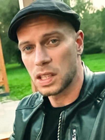 A close-up, chest-up shot shows a man wearing a black flat cap and a black leather jacket. He has fair skin, blue eyes, and short-cropped hair, with some stubble on his chin and jawline. His mouth is slightly open, revealing teeth. The background is blurred, showing greenery, suggesting an outdoor setting. There are no visible indications of the city of Dondușeni, Moldova.