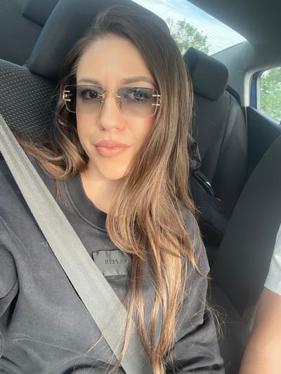 A woman with long brown hair and sunglasses lies reclined in a car seat, wearing a black shirt with a gray seatbelt across her. The scene is inside a car, with the car seat and other interior parts visible. The location, Shishmantsi, Bulgaria, is not identifiable from the image alone.