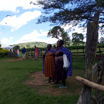 The image shows two women in traditional Maasai attire standing on a grassy area in Keyian ward, Kenya. They are wearing colorful beaded necklaces and blue shawls. One woman wears a brown dress, while the other wears a purple dress. To the left, other people are visible near a fence and a white tent. The background includes a green, hilly landscape under a blue sky with scattered clouds. Three birds are flying in the sky. In the foreground, a weathered wooden fence post is visible.