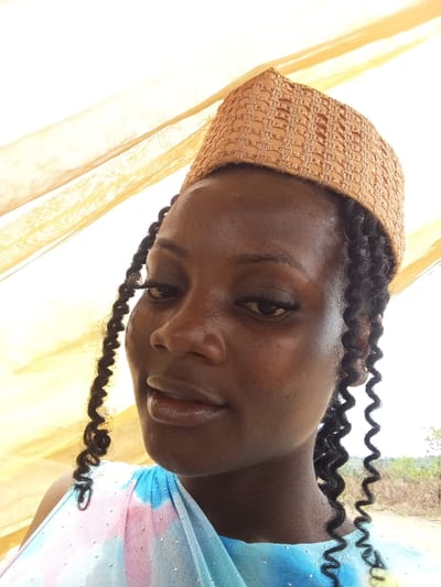 A female subject is depicted from the chest up, located in Alayi, Nigeria. She has dark skin, dark curly hair styled in multiple thin braids, and wears a light brown, woven cap with a grid pattern on her head. Her attire includes a garment featuring blue, pink, and white tie-dye-like patterns, possibly of sheer fabric, adorned with small reflective dots. She has prominent eyelashes and small, round, light-colored stud earrings. The subject is looking directly towards the camera with a slight smile. The immediate background shows a translucent, yellowish-orange fabric drape. The distant background on the right reveals an outdoor area with dry vegetation under a bright sky. The scene is illuminated by daylight.
