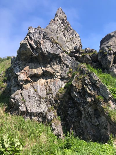 The image shows a steep, rocky hillside with large boulders scattered across it. Green grass and some small yellow flowers grow between the rocks and on the slopes. The sky is clear and blue. The scene is an outdoor natural environment, likely a mountainous or hilly area. There are no people or structures visible in the image. The location is described as Ola, Russia, implying this is a natural landscape within that region.