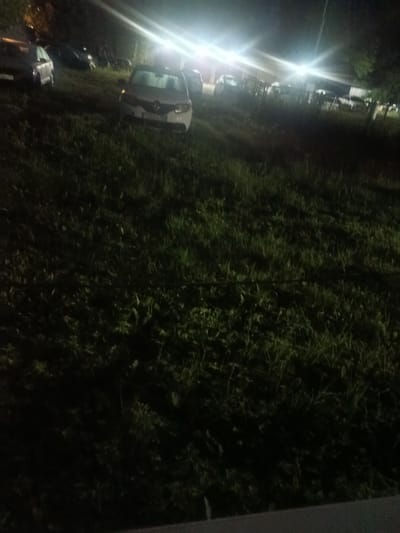 The image shows a dark, grassy area illuminated by bright artificial lights. Several cars are parked in the grass. In the foreground, a white car is visible, facing the camera. Its license plate is partially obscured. To the left, a darker colored car is also visible, parked at an angle. In the background, more vehicles are present, along with some structures that appear to be buildings or fences, also illuminated by the lights. The overall impression is of a parking area or an informal gathering space at night, possibly in Mikhnevo, Russia, given the mention of the location. There are no people visible in the image. The grass is tall and somewhat overgrown, with shadows cast by the artificial lights.