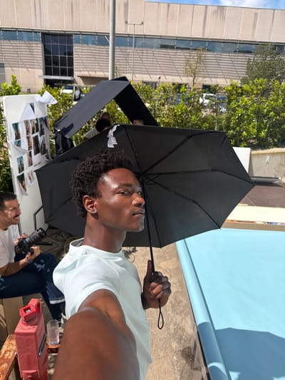 The image depicts an outdoor photoshoot in a city. A young, dark-skinned man holds a black umbrella over himself, ostensibly for shade, as he looks upward. He wears a light-colored t-shirt. A person partially visible at the bottom of the frame holds a camera, appearing to take a photograph. In the background, there is a building, trees, a light reflector, a blue backdrop, and a table covered with printed photos. Based on the prompt, the city is Fibracolor, Spain.