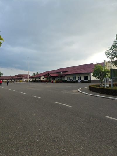 In Glokaron, Indonesia, the image shows an exterior view of a building with a red tiled roof and a beige facade. The building appears to be a public facility, possibly an airport or a civic center, with multiple sections and a long, covered walkway. The foreground features an asphalt road with white dashed lines, suggesting a parking area or access road. Two individuals are walking on the road in the middle ground, one wearing a red shirt and the other a dark shirt. Several trees and manicured hedges are visible around the building and the periphery of the paved area. The sky is overcast with grey clouds, and the overall lighting suggests it is late afternoon or early morning.